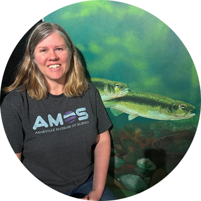 Image of Heath with shoulder length hair smiling wearing an AMOS shirt sitting by trout painting.