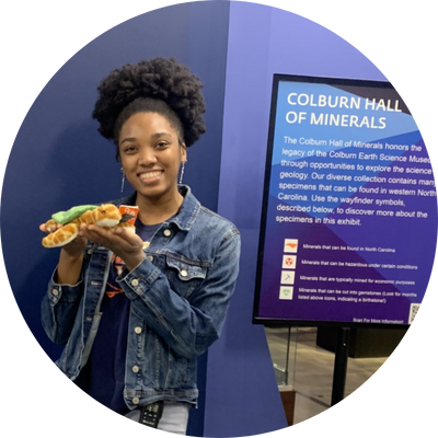 Keyana smiles and holds a plush turtle next to the Colburn Hall of Minerals sign